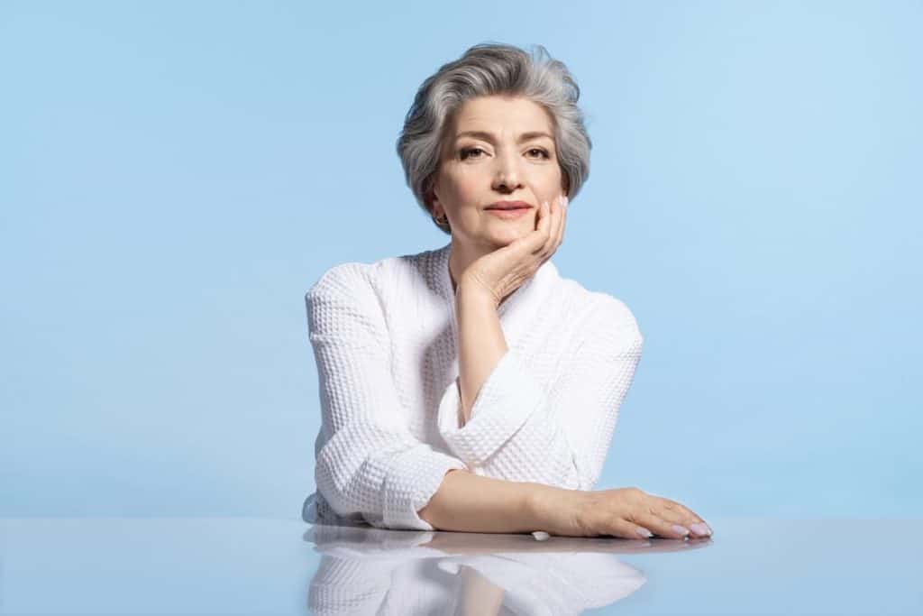 A short haired mature woman with a gray blended bixie haircut, sitting. Isolated on a blue background