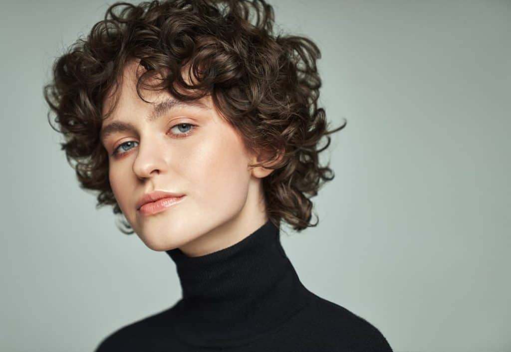 A curly and short-haired woman wearing makeup and black turtle neck isolated on a gray background