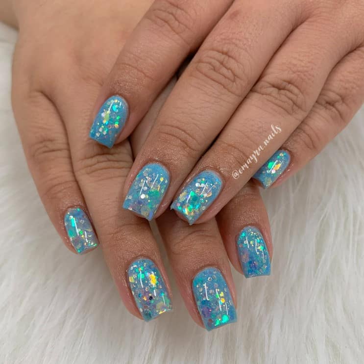 A closeup of a woman's hands that has a mid-length square nails with aquamarine nail polish and oversized glitter flakes