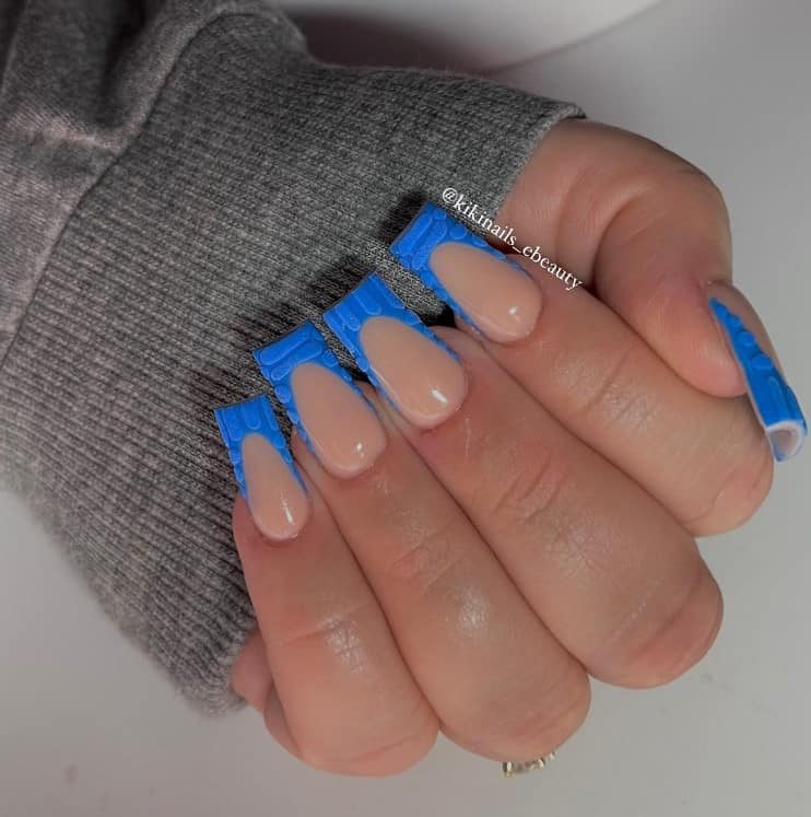 A closeup of a woman's hand with a glossy nude nail polish that has blue tips with textured croc-print designs
