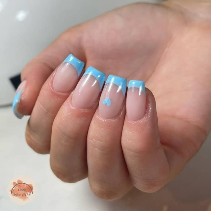 A closeup of a woman's hand with nude nail polish base that has a sky blue French tips and a a lone heart on select nail