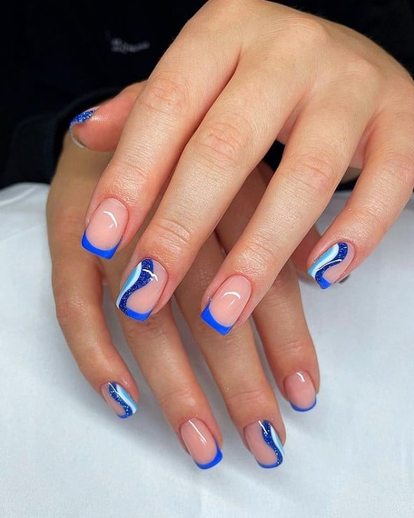 A closeup of a woman's hands with a glossy peach nail polish that has sideway waves in different shades of blue on select nails