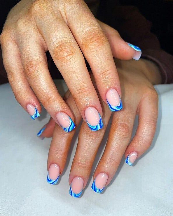 A closeup of a woman's hands with nude nail polish base that has French tips with swirls in different shades of blue