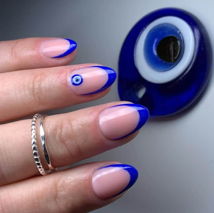 A closeup of a woman's hand with a nude nail polish base that has U-tips in blue  and Nazar nail art on select nail