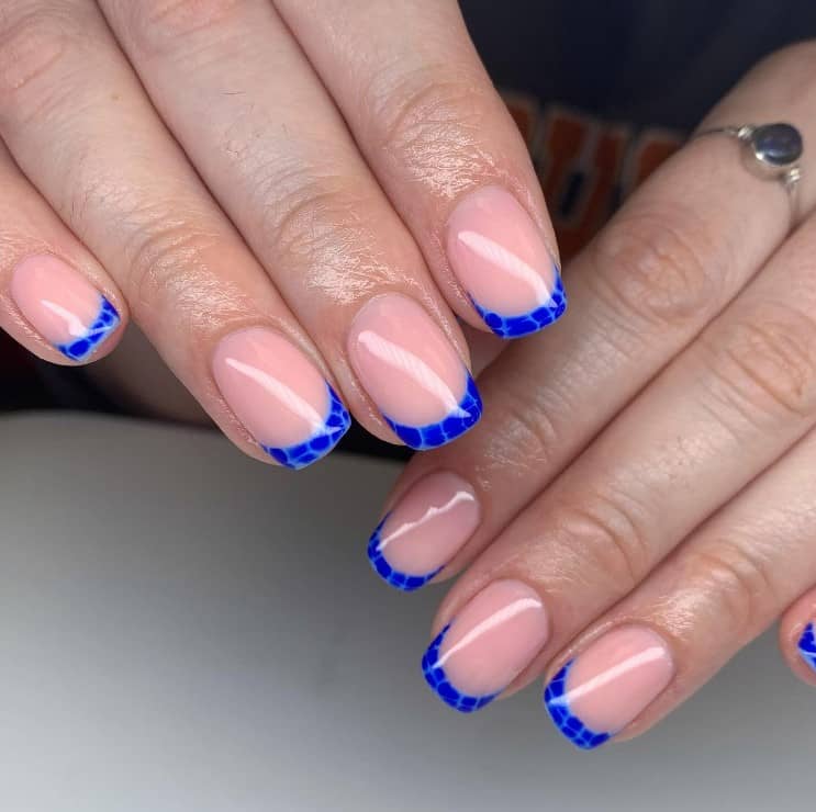 A closeup of a woman's hands with a glossy peach nail polish base that has thin blue French tips with dark blue animal print  