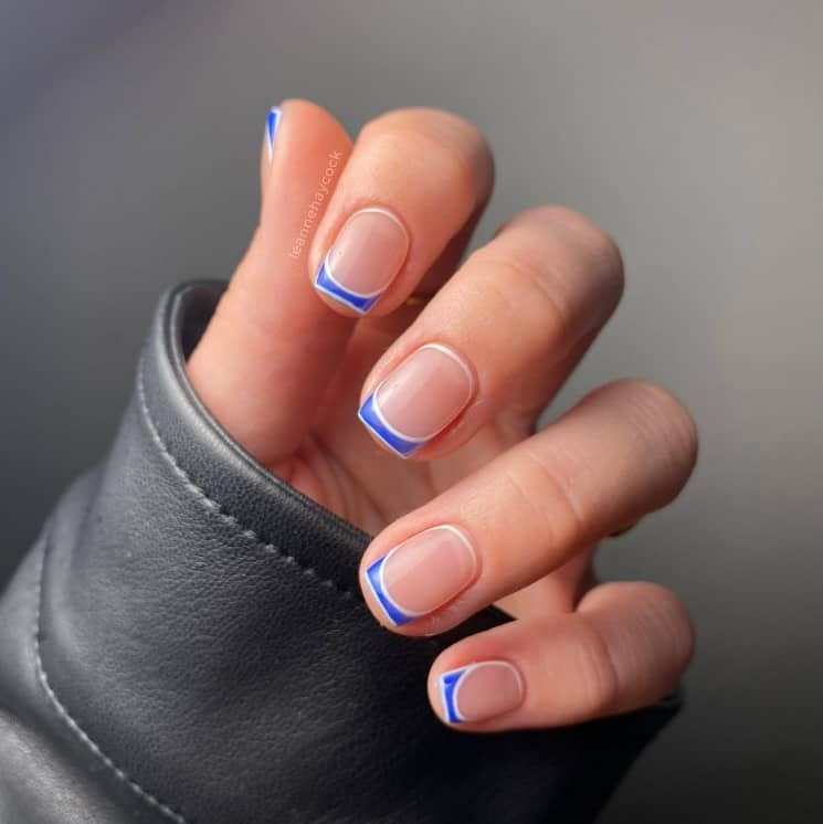 A closeup of a woman's hand with nude nail polish that has mini blue French tips and edges. Tips outlined with white nail polish