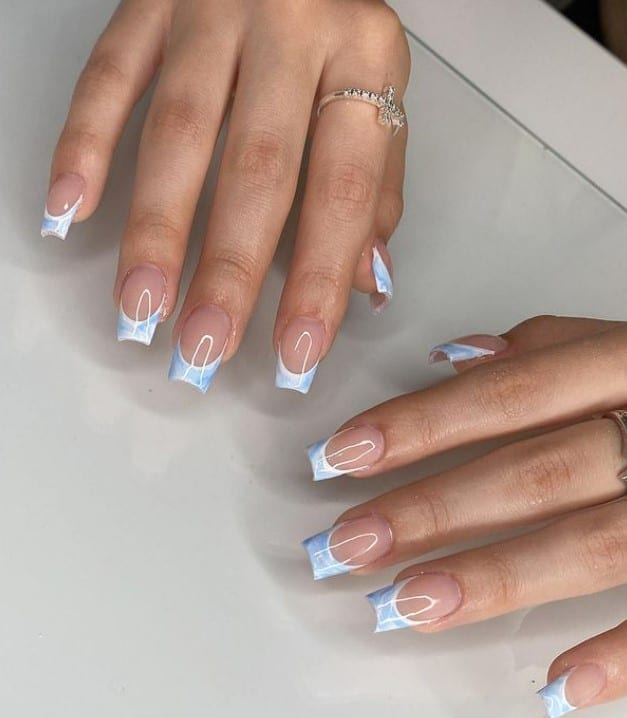 A closeup of a woman's hands with a glossy nude nail polish base that has white-and-light-blue marble tips on long square nails