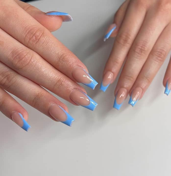 A closeup of a woman's hands with a glossy nude nail polish that has sky blue French tips on long coffin nails