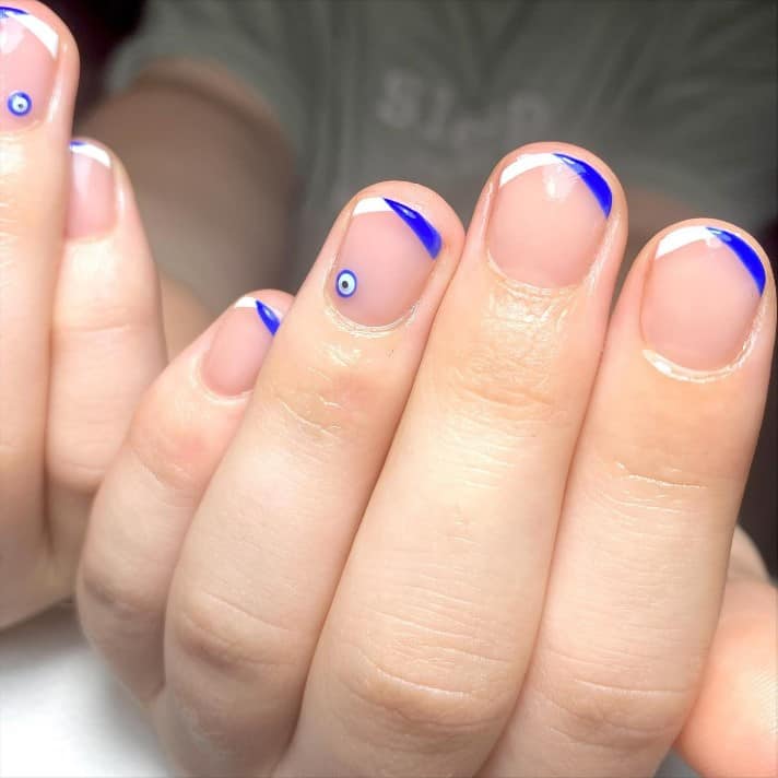 A closeup of a woman's hands with pale pink nail polish that has diagonal V-tips in white and blue on nails and mysterious Turkish eye pendant nail art