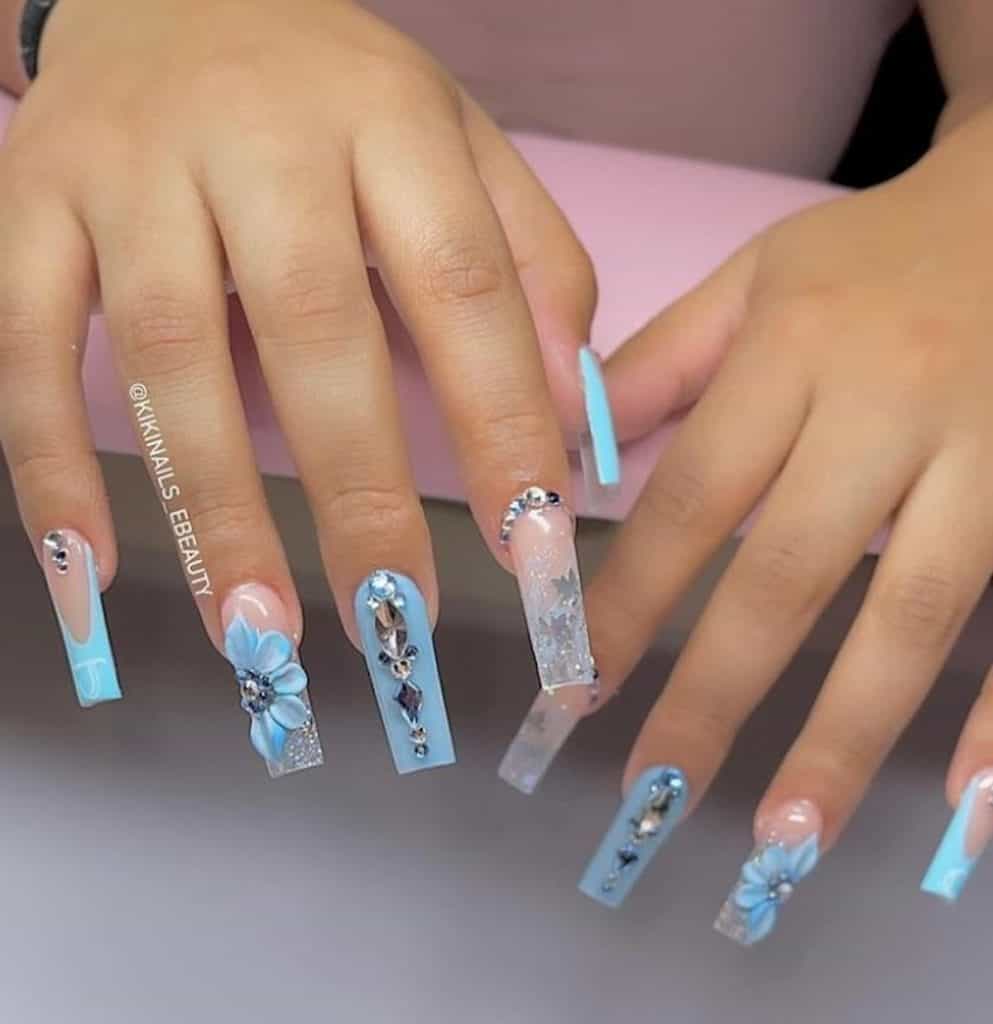 A closeup of a woman's hands with sky blue nail polish that has glitter, diamonds, and 3D flowers nail designs