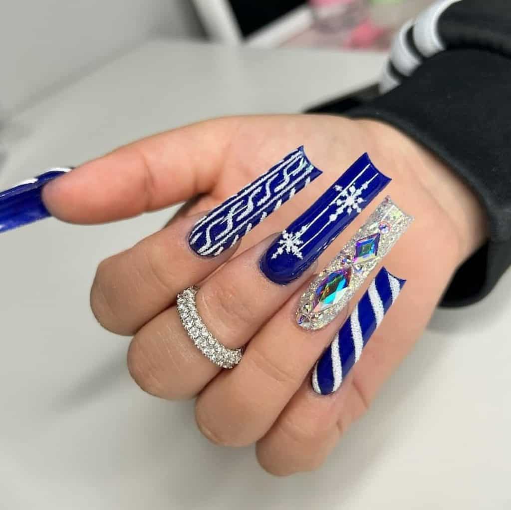 A closeup of a woman's hand with a blue nail polish that has winter-inspired designs in white glitter shine, glittery silver polish and accessorized with iridescent diamonds