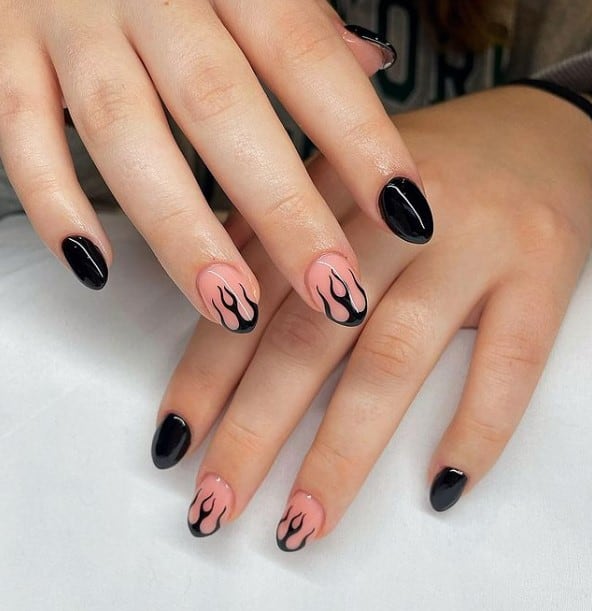 A closeup of a woman's hands with a combination of glossy nude and black nail polish that has black flame design nail art on select nails