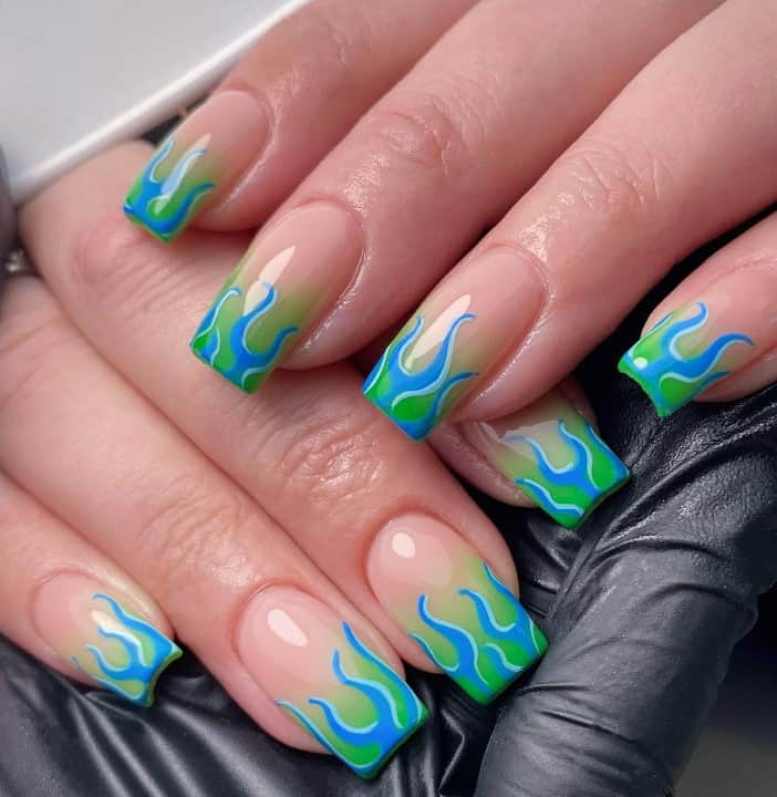 A closeup of a woman's hands with a glossy nude nail polish that has blue flames outlined by a lighter blue and green ombre