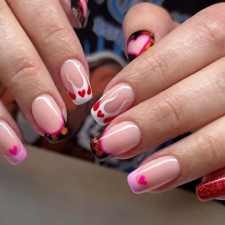 A closeup of a woman's hands with a glossy nude nail polish that has white flame nails complemented with pink and red hearts, amber French tips, and red glitter
