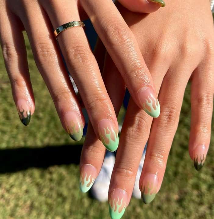 A closeup of a woman's hands with a nude nail polish base that has a flame art in several shades of green on the tips