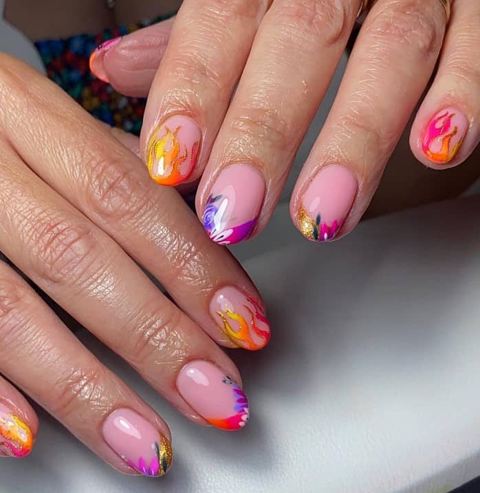 A closeup of a woman's hand with a glossy pale pink nail polish that has yellow, pink, and orange gradient flames nail art with gold glitter accents