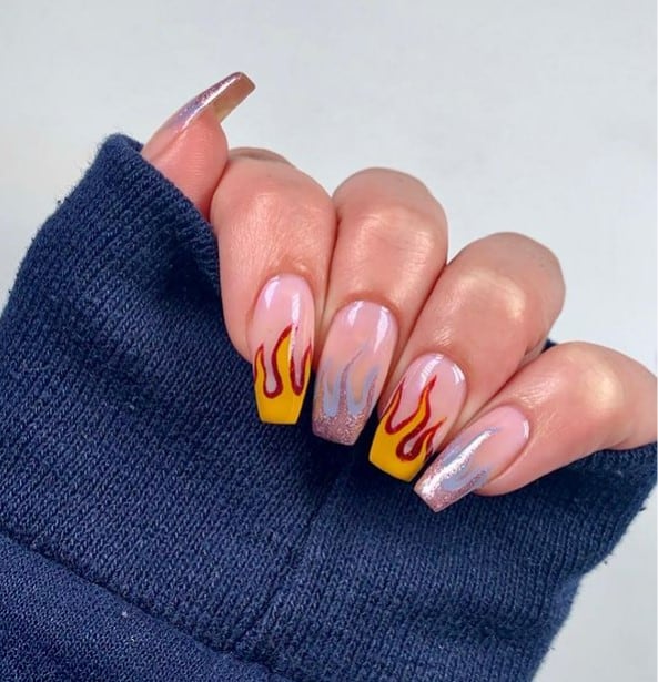 A closeup of a woman's hand with nude nail polish that has cute yellow fire nails outlined with red and alternated with silver fire nails outlined with icy blue are for you