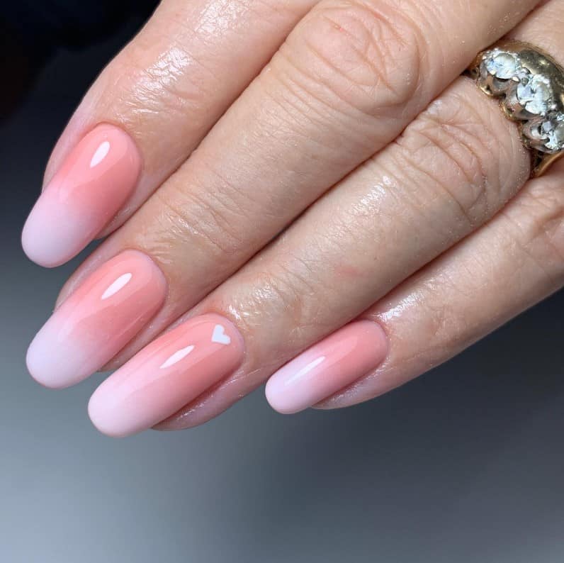 A closeup of a woman's hand with a peach French ombré nails and gradient of pink nail tips that has small white heart on select nails 