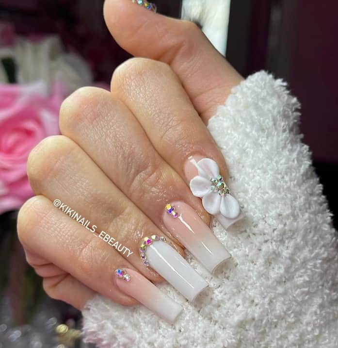 A closeup of a woman's hand with nude nail polish that has white nail tips multicolored rhinestones and half-a-flower 3D design 