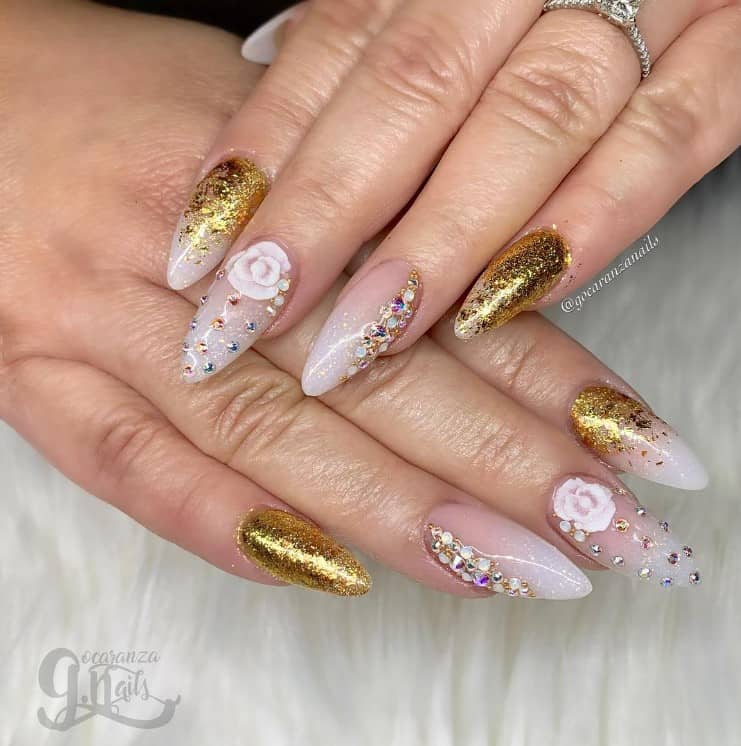 A closeup of a woman's hands with a combination of nude and gold glitter nail polish that has dainty white flowers, colorful rhinestones, and white glitter