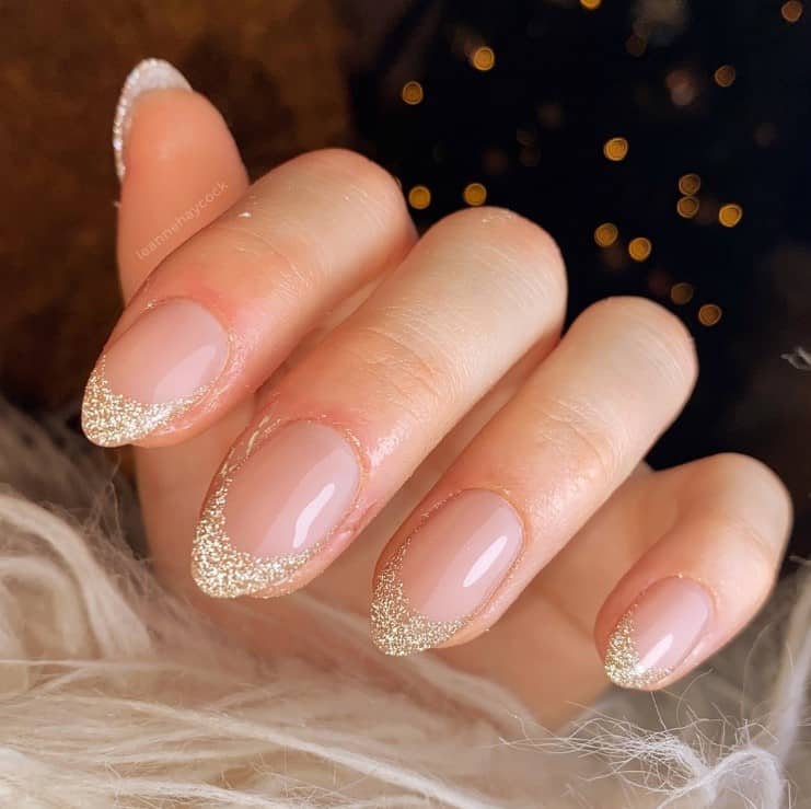 A closeup of a woman's hand with glossy nude nail polish that has glittering gold French tips