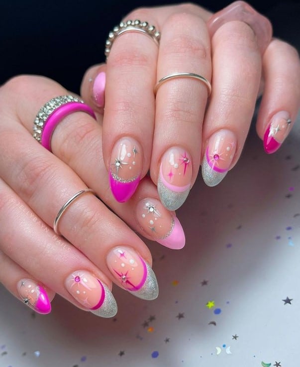 A closeup of a woman's hands with nude nail polish base that has multi-colored nail design with alternating pink and silver glitter tips, silver gems and chrome silver stars