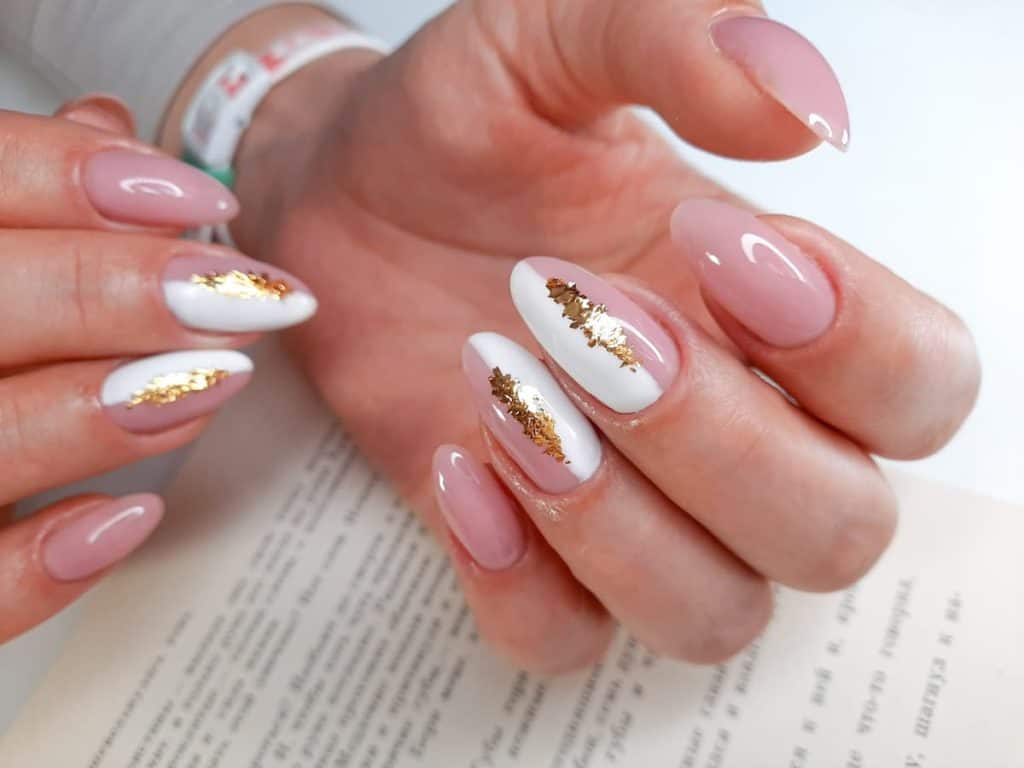 A closeup of a woman's fingernails with a pink nail polish and half-and-half white and pink nail polish design on select nails that has a splash of gold foil on select nails