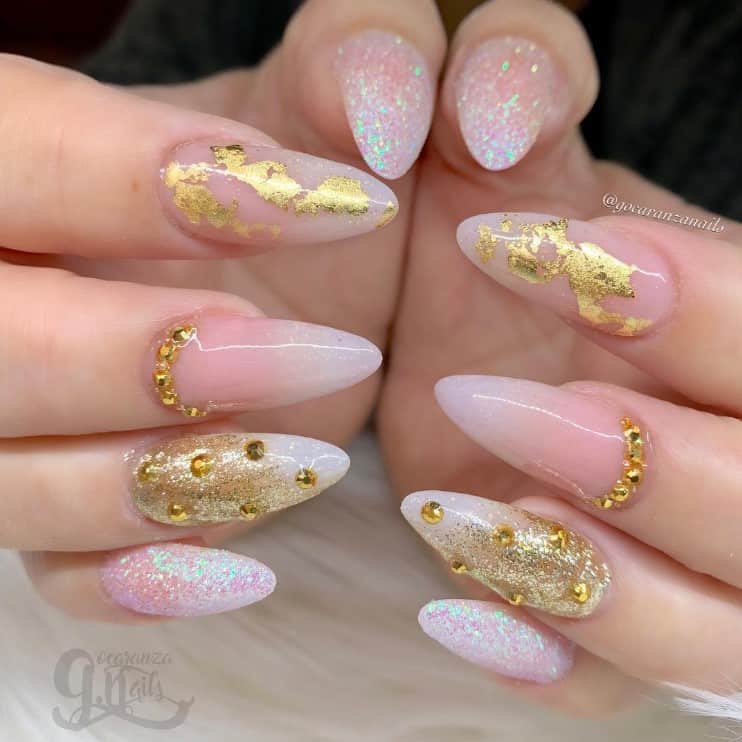 A closeup of a woman's fingernails with a glossy baby pink nail polish that has white tips, gold rhinestones, gold foil, and gold glitter