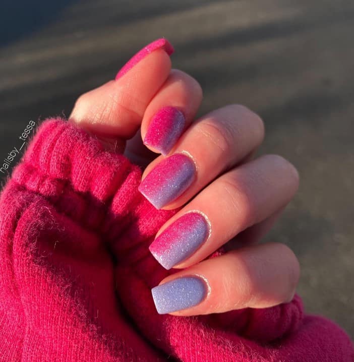 A closeup of a woman's fingernails with a combination of pink and purple nail polish that has a glitter finish and a vertical ombre design that transitions from pink to purple