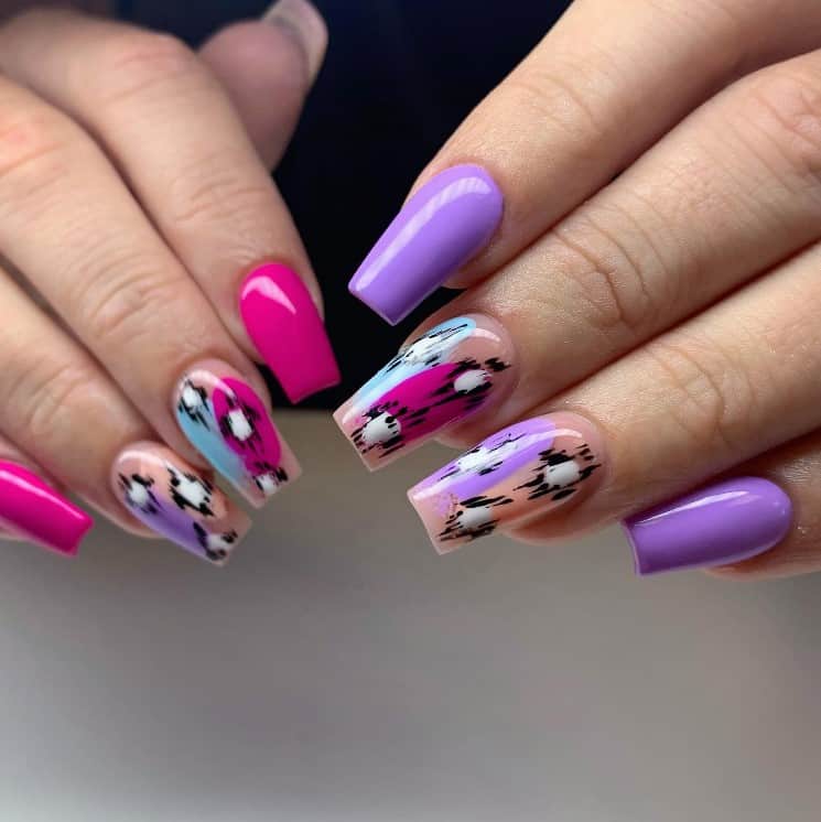 A closeup of a woman's fingernails with a nude, pink and purple nail polish that has irregularly shaped circles in pink, lavender, light blue, and white, as well as the black strokes 