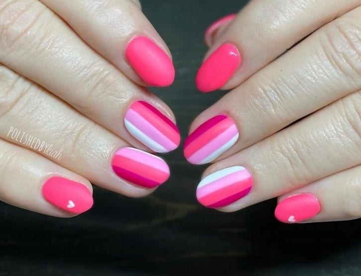 A closeup of a woman's hands with a pink nail polish that has different shades of pink gel polish and white polish vertical stripes and solid with a tiny heart