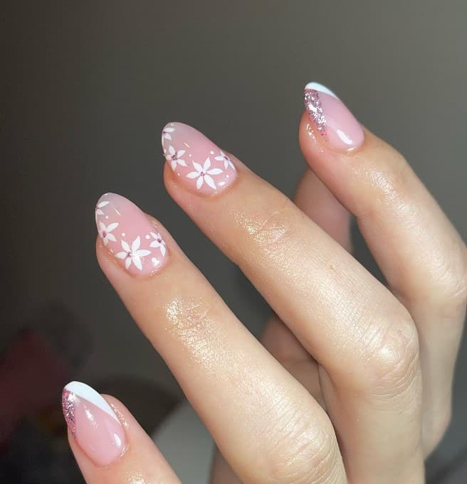 A closeup of a woman's hand with light nude pink nail polish that has dainty white flowers and pink glitter