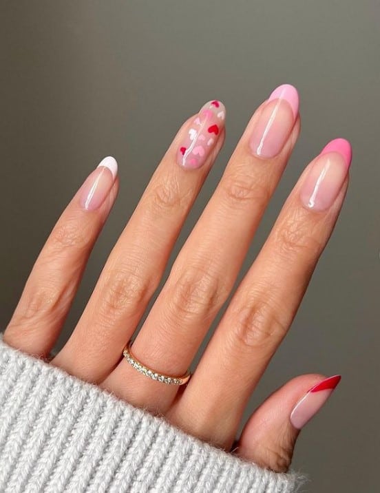 A closeup of a woman's hand with glossy nude nail polish base that has French tips in red, white, and different shades of pink, and an accent nail that’s loaded with adorable little hearts 
