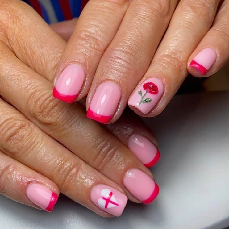 A closeup of a woman's hands with pastel pink nail polish base that has hot pink tips and a hand-painted red rose and a white heart with a red cross in the middle