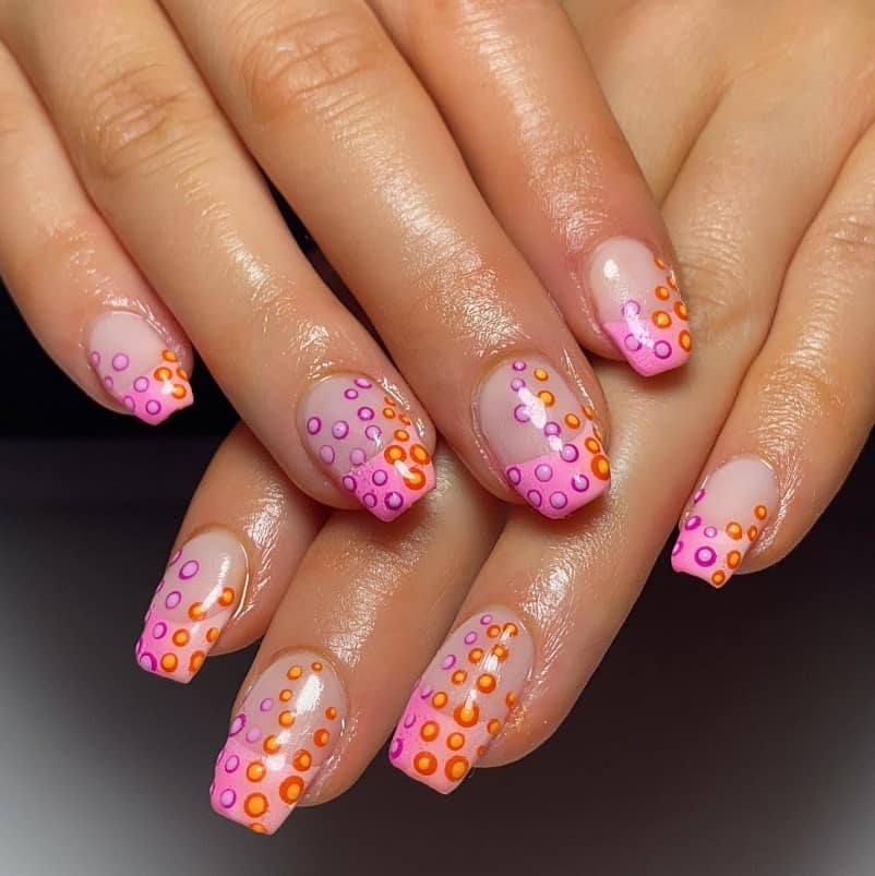 A closeup of a woman's hands with nude nail polish base that has pink tips, funky orange and purple dots arranged in irregular patterns