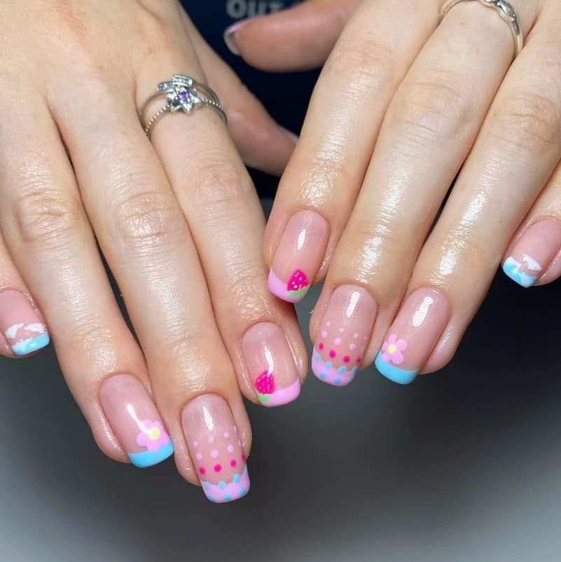 A closeup of a woman's hands with pale pink nail polish base that has  pink and blue French tips, strawberries, clouds, polka dots, and flowers nail designs