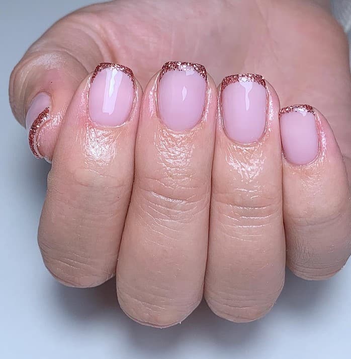A closeup of a woman's hand with nude pink nail polish base that has rose gold glitter tips