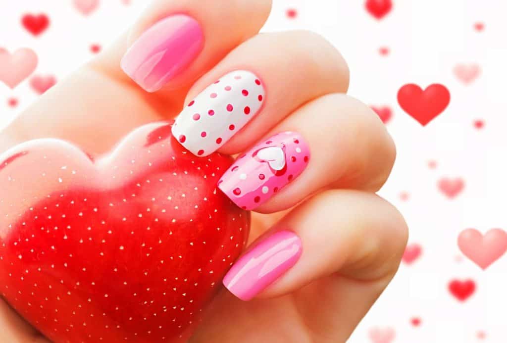 A hand of a woman with white and pink colored nails with heart designs while holding a heart