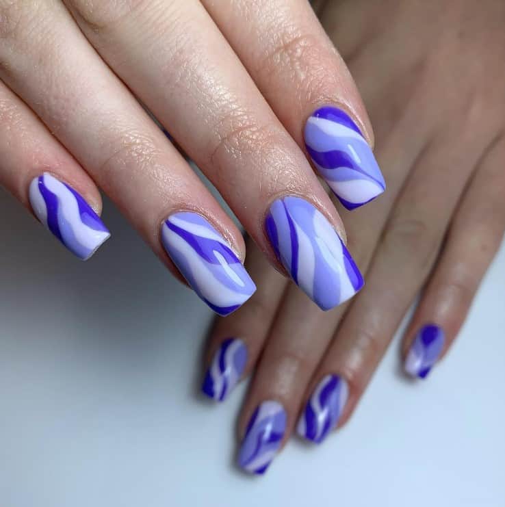 A closeup of a woman's fingernails with bluish-purple nail polish that has white swirls nail designs 