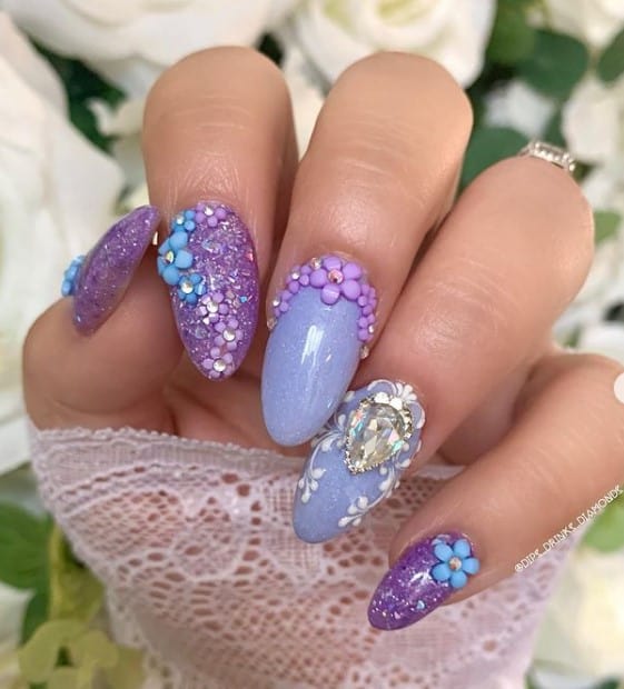 A closeup of a woman's fingernails with lighter and darker purple nail polish that has rhinestones and 3D flowers nail designs