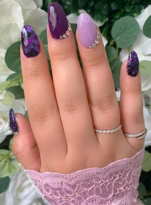 A closeup of a woman's fingernails with different shades of purple nail polish that has glitter and rhinestones