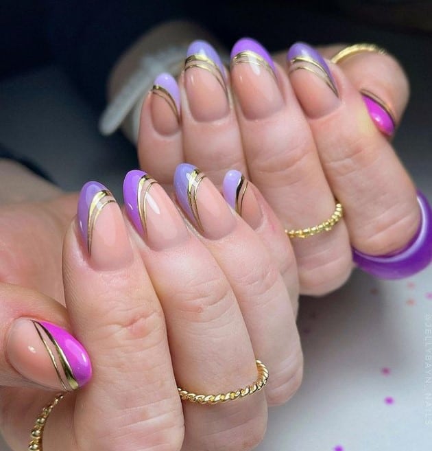 A closeup of a woman's fingernails with nude nail polish that has different shades of purple nail tips and gold outlines