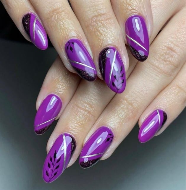 A closeup of a woman's fingernails with a glossy dark purple nail polish that has black leaves, thick glittery black tips, and thin silver lines