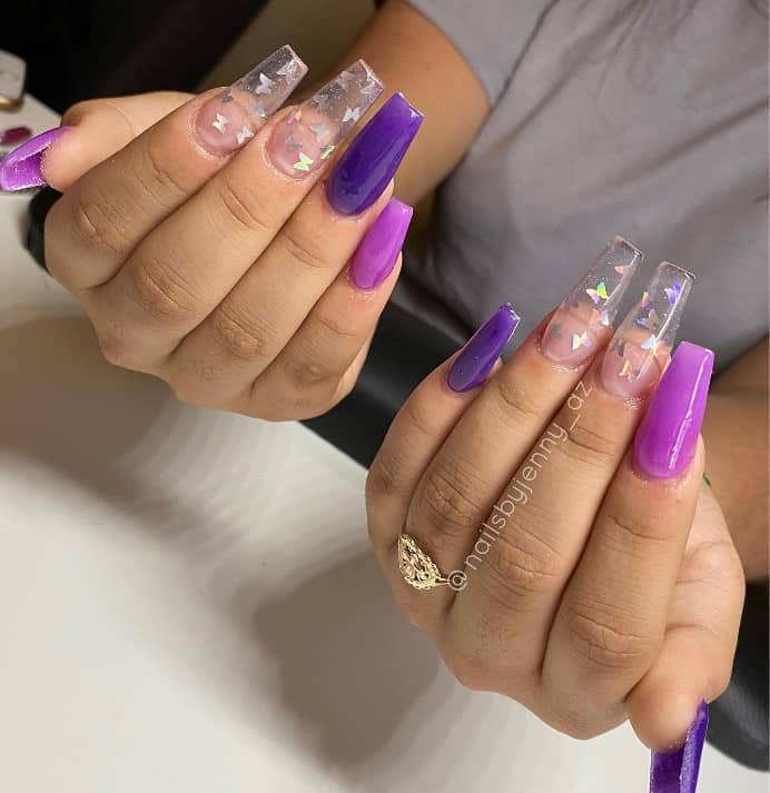 A closeup of a woman's fingernails with different shades of purple nail polish that has light and dark purple polish that has cute little butterfly stickers nail designs