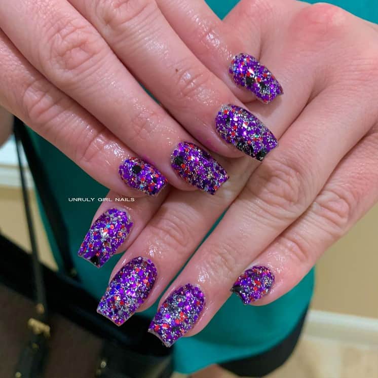 A closeup of a woman's hands with purple glitter nail polish that has red and black flecks