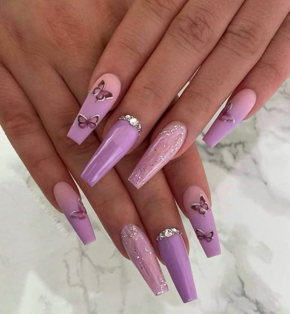 A closeup of a woman's fingernails with pink that fades into lilac nail polish that has butterfly decals or hand-painted nail designs 