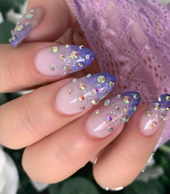 A closeup of a woman's fingernails with a purple ombré gradient effect nail polish that has rhinestones and subtle glitter