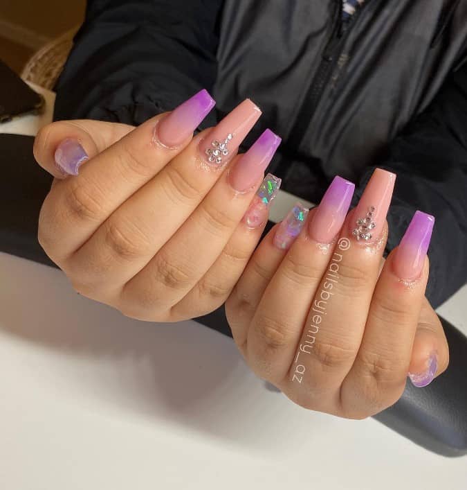 A woman's beautiful long fingernails with  pink and purple ombré nail polish that has rhinestones and a clear yet holographic accent nail