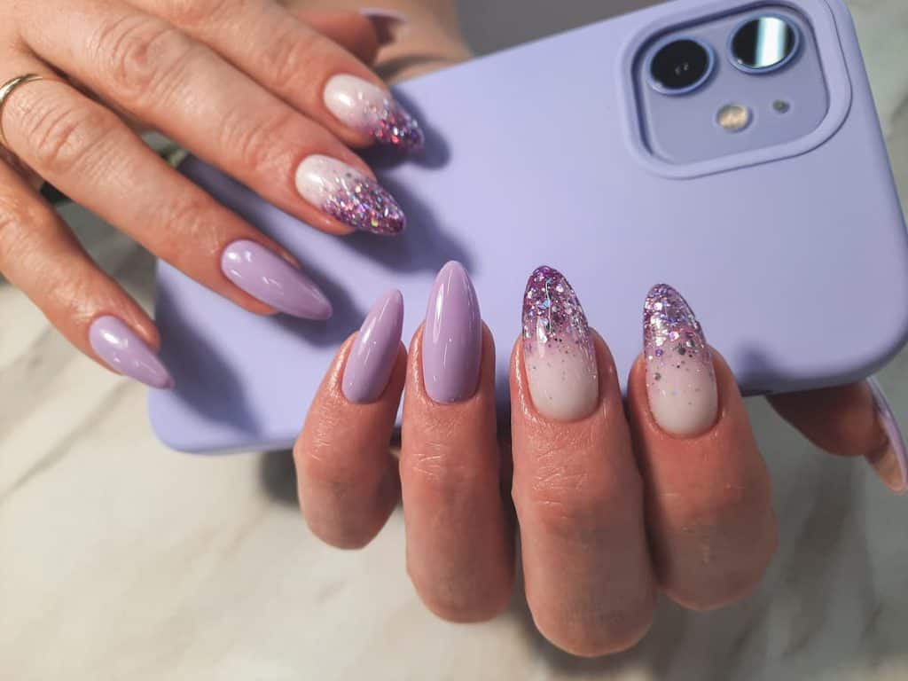 A closeup of a woman's fingernails with a glossy purple and white nail polish that has a gradient effect and chunky glitter on select nails
