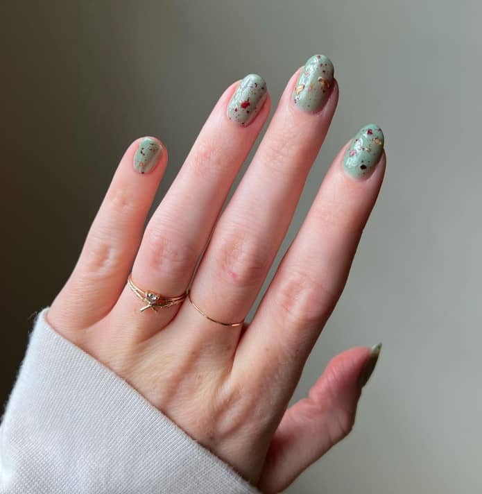 A woman's oval or egg-shaped fingernails with sage green nail polish that has different colors for the speckles nail designs